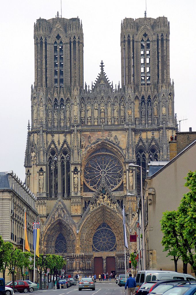 A richly carved twin-towered cathedral façade closes the view along a street lined with buildings, trees, parked cars and pedestrians.