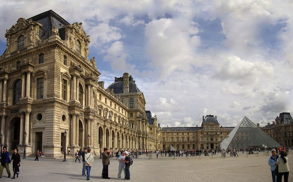 A long richly ornamented palace range with paired columns and projecting pavilions borders a broad court beside a modern glass pyramid.
