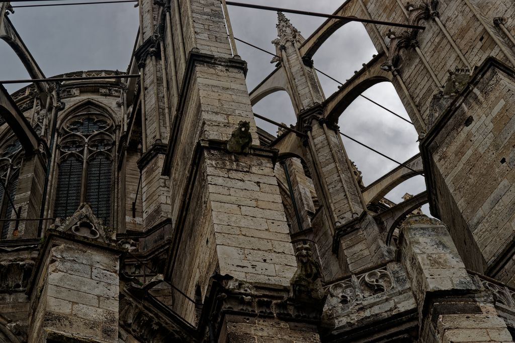 An upward view shows several stone flying buttresses and gargoyles between tall masonry masses, with dark metal rods spanning the gaps.