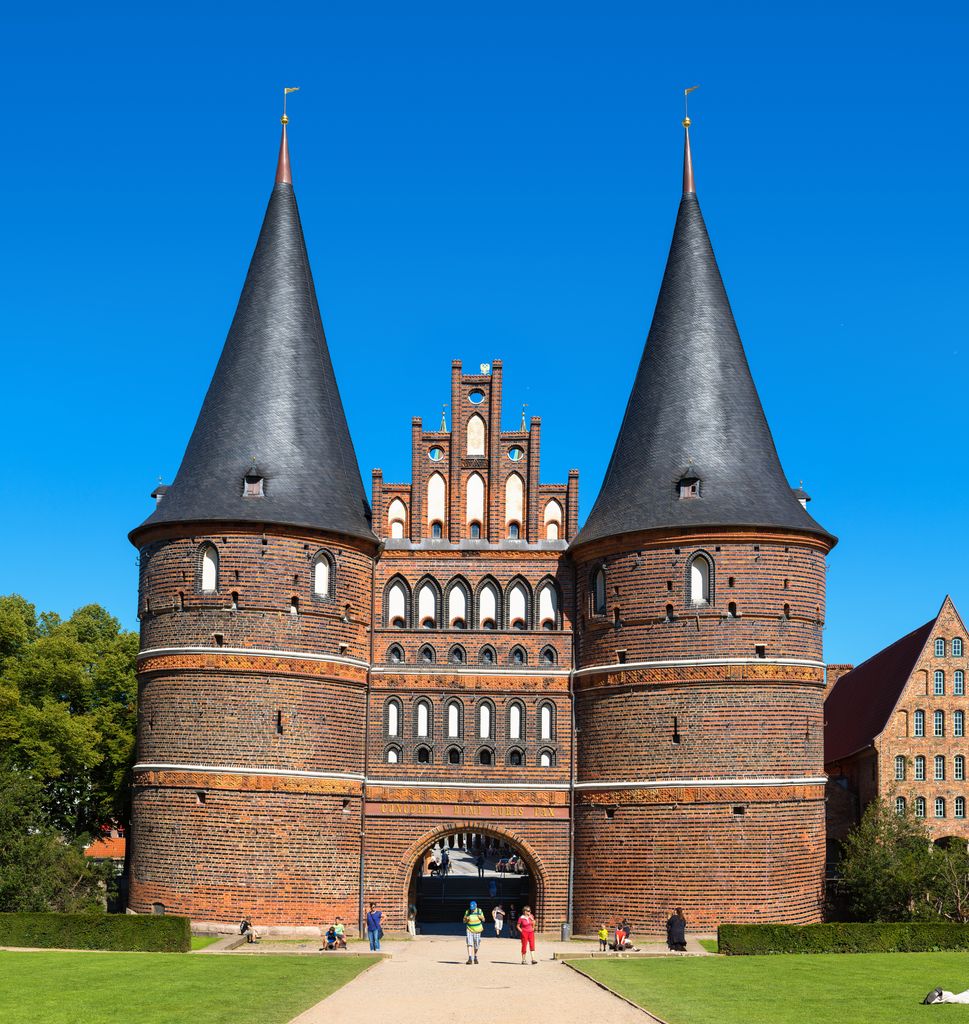 Two round brick towers with tall dark conical roofs flank a stepped central façade pierced by pointed openings above a gateway.