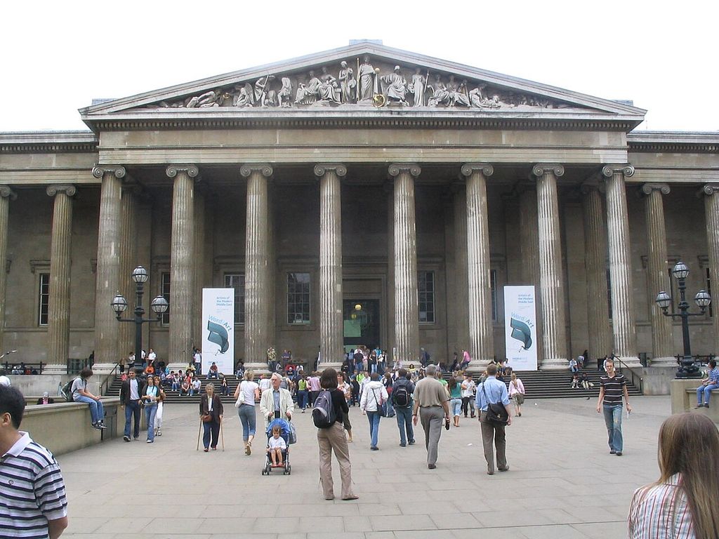 A low triangular sculpted pediment crowns a deep stone colonnade above a crowded museum entrance court.