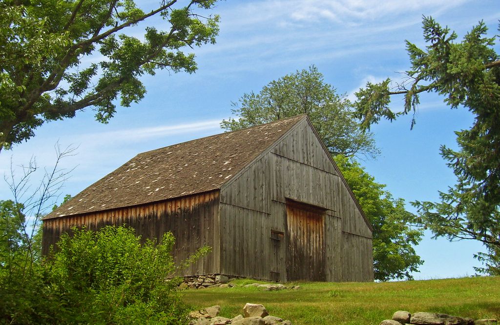 A weathered timber barn has a broad pitched roof, a plain boarded gable end and large central wagon doors on a grassy rise.