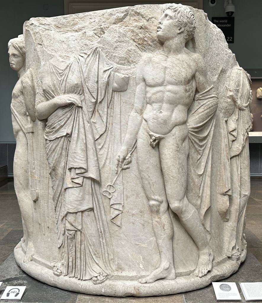 A large round marble column drum carved with high-relief figures stands in a museum gallery.