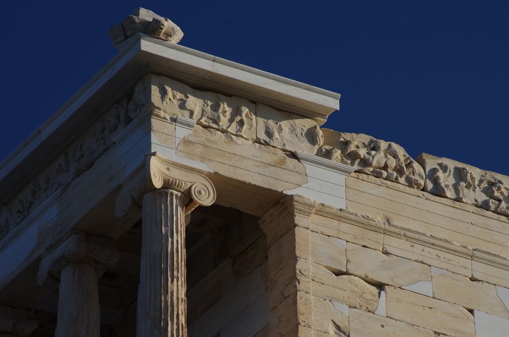 An outward-facing volute capital supports carved horizontal bands and a continuous figure frieze above a fluted column.