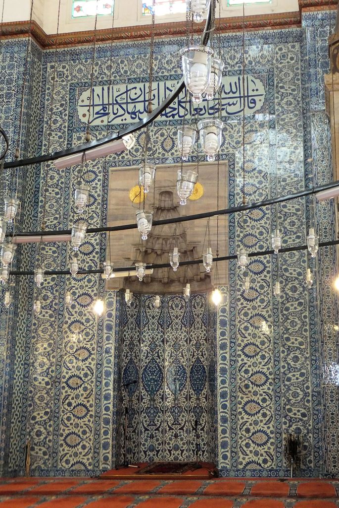 Dense blue-and-white floral tiles and an Arabic inscription surround a stone mihrab beneath rows of hanging glass lamps.