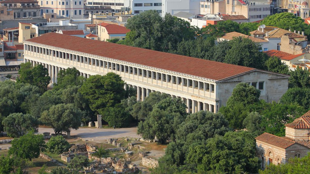 A long two-storey colonnaded building with a red tiled roof stands among trees and archaeological remains below modern Athens.