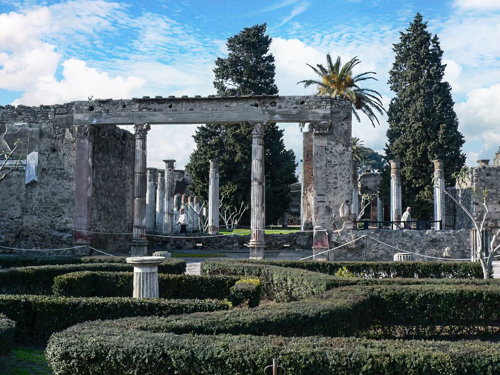 Incomplete column rows and masonry openings surround a planted garden with hedges, ropes and several visitors at Pompeii.