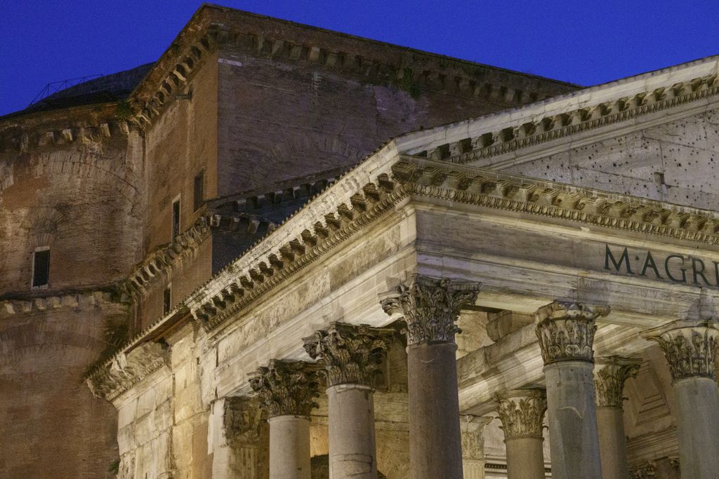 Illuminated Corinthian columns and a pale pediment cross in front of a dark cylindrical brick wall and stepped dome under a deep blue evening sky.