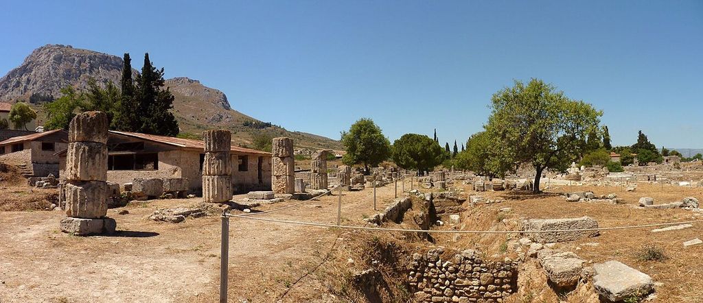 Weathered column drums and low excavated walls extend across a dry archaeological site below a rocky mountain.