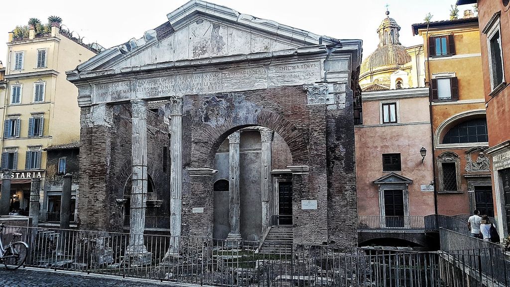 Two weathered marble columns flank a tall brick arch beneath a pale inscribed upper front, closely surrounded by later city buildings.