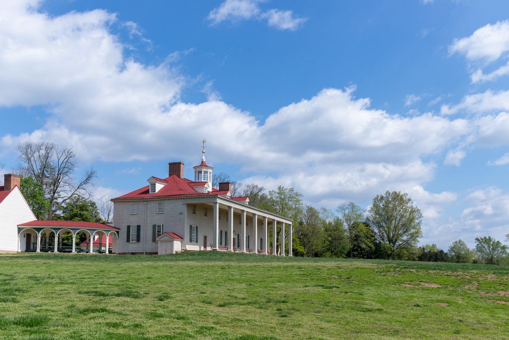 Tall square posts carry a long roof across a white mansion facing a broad lawn, with a lower arched passage extending at left.