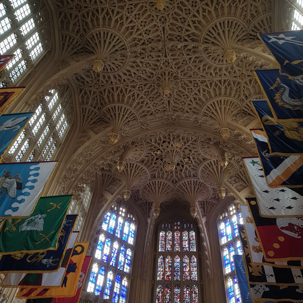 Dense stone fans, hanging pendants and lace-like tracery cover a chapel ceiling above bright windows and rows of colourful banners.