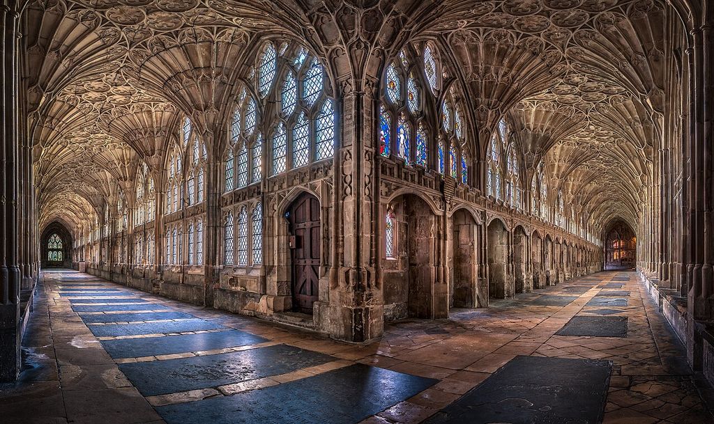 Two long stone cloister passages meet beneath dense fan-shaped vaults beside large traceried windows.