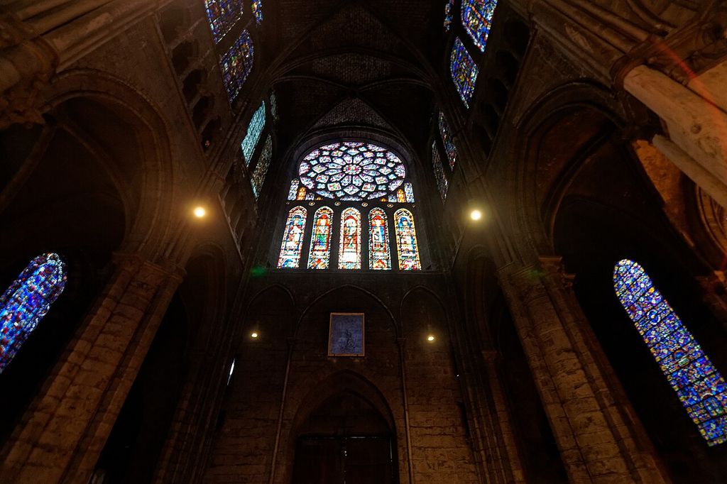 A luminous circular stained-glass rose sits above five tall lancets within a shadowed stone transept wall.