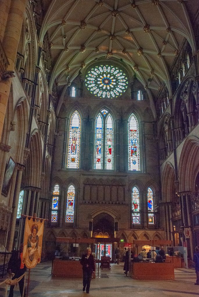 A small luminous rose and three tall lancets rise in the end wall of a vaulted transept above visitors and church furnishings.