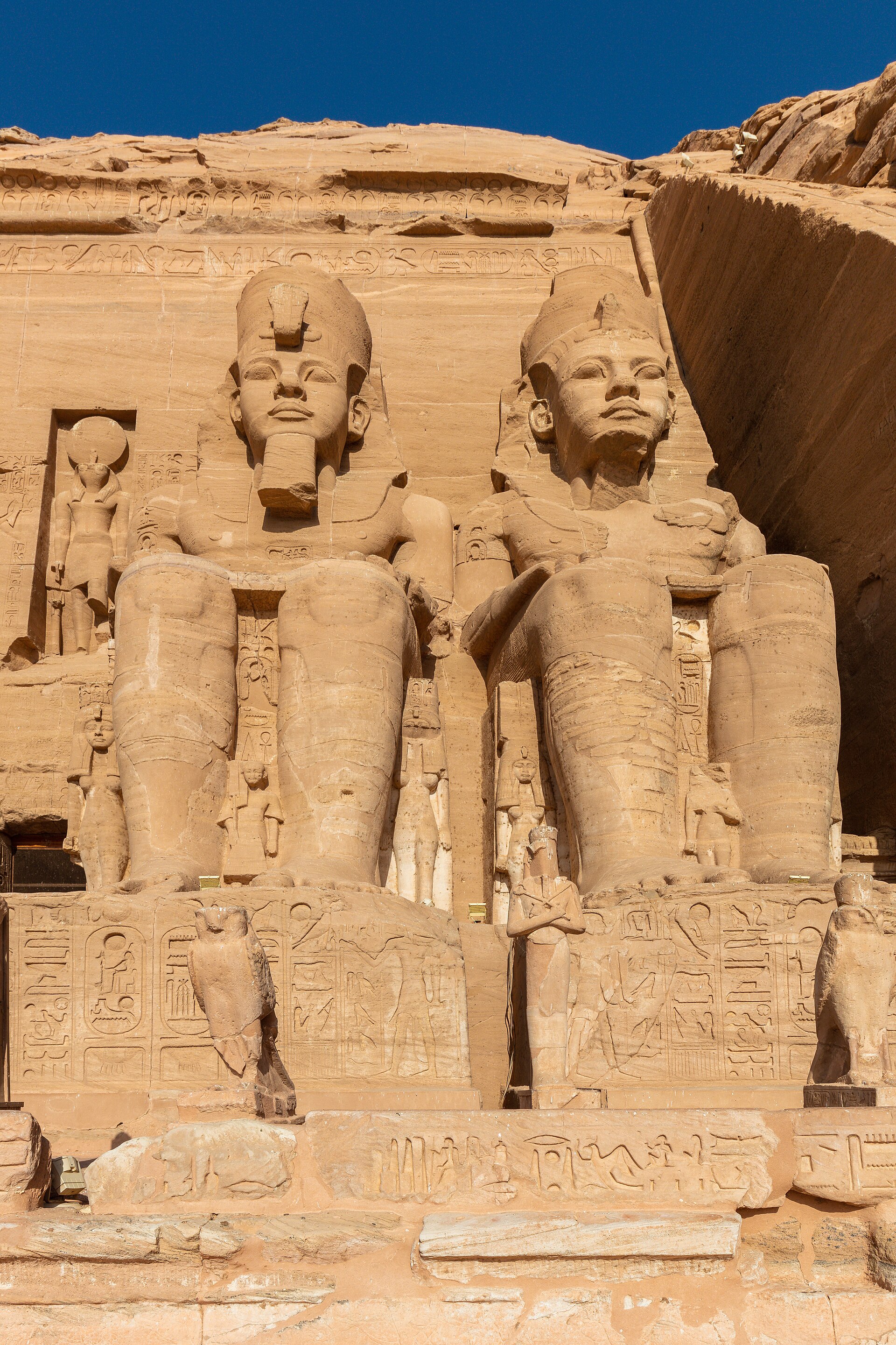 Close view of two seated Ramesses colossi at the Great Temple, with smaller carved family figures at their legs and the baboon frieze and portal niche above.