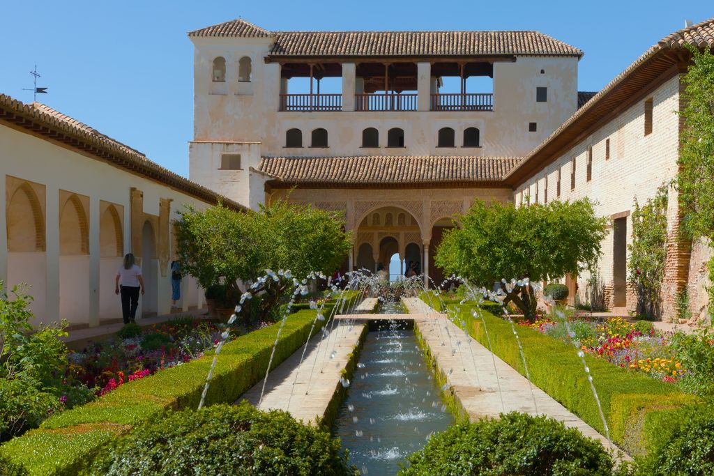 A long garden channel runs between green hedges and flowers as crossing fountain jets lead toward a white arcaded pavilion at the Generalife.