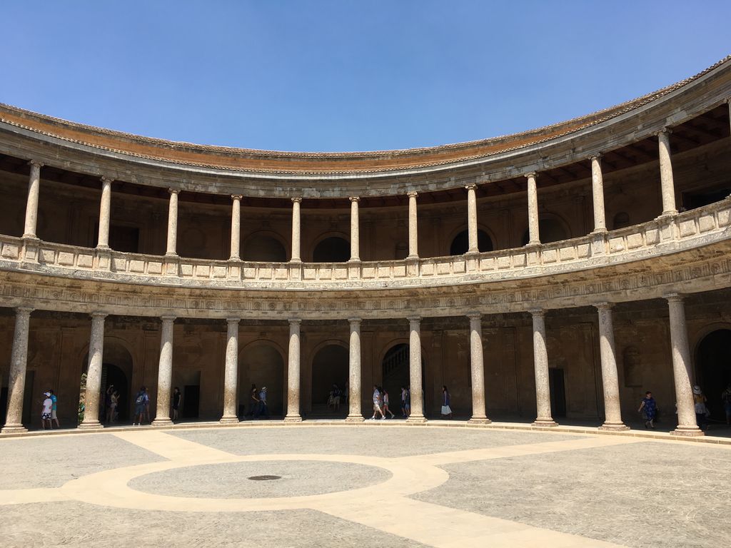 Two storeys of curving stone colonnades surround a broad circular open courtyard beneath a clear blue sky.