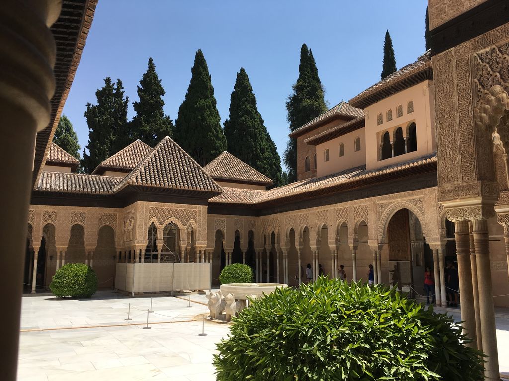 A sunlit court is enclosed by low tiled roofs and arcades of slender columns, with the lion fountain partly visible behind plants and barriers.