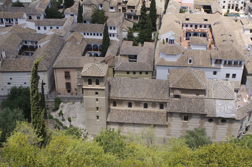 A steep elevated view looks down across closely packed tiled roofs, small courtyards, galleries, towers and garden trees in part of the Alhambra.