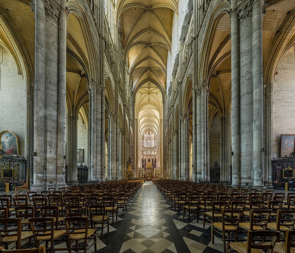 A long cathedral nave has clustered stone piers, tall pointed arcades, narrow upper stages, high windows and repeated rib vaults above rows of chairs.