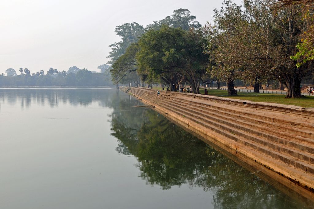 A long stepped sandstone embankment runs beside calm moat water that reflects trees, with a few visitors on the shaded bank.