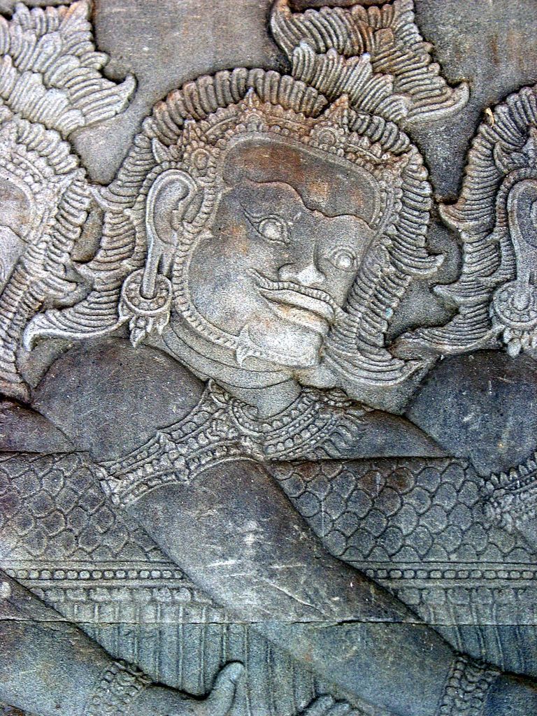 A close stone relief shows an ornate asura gripping a thick serpent while rows of hands and neighbouring figures continue beyond the frame.
