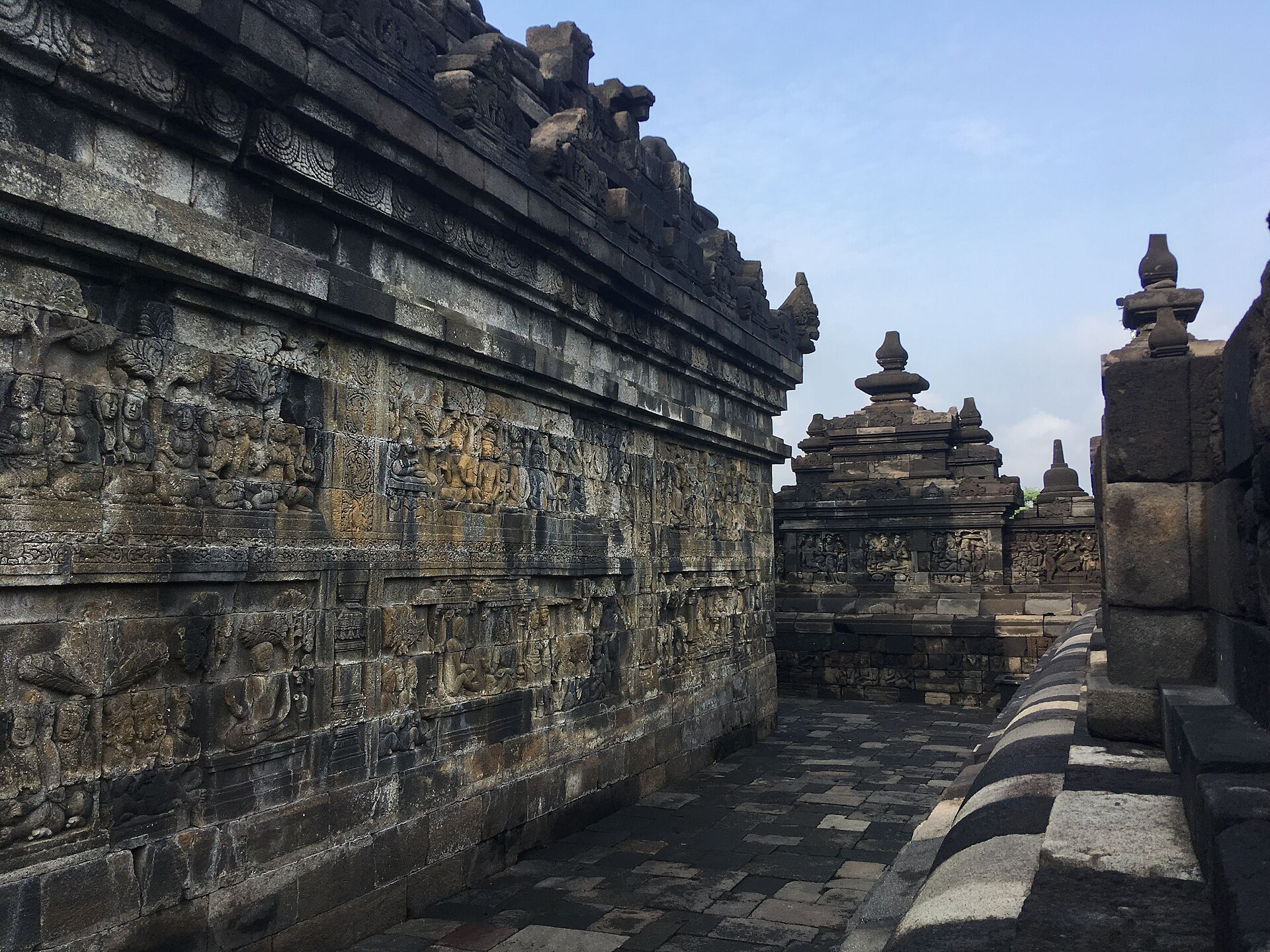 Roofless carved Borobudur gallery with relief-bearing inner wall, opposite balustrade, stone paving and open sky.