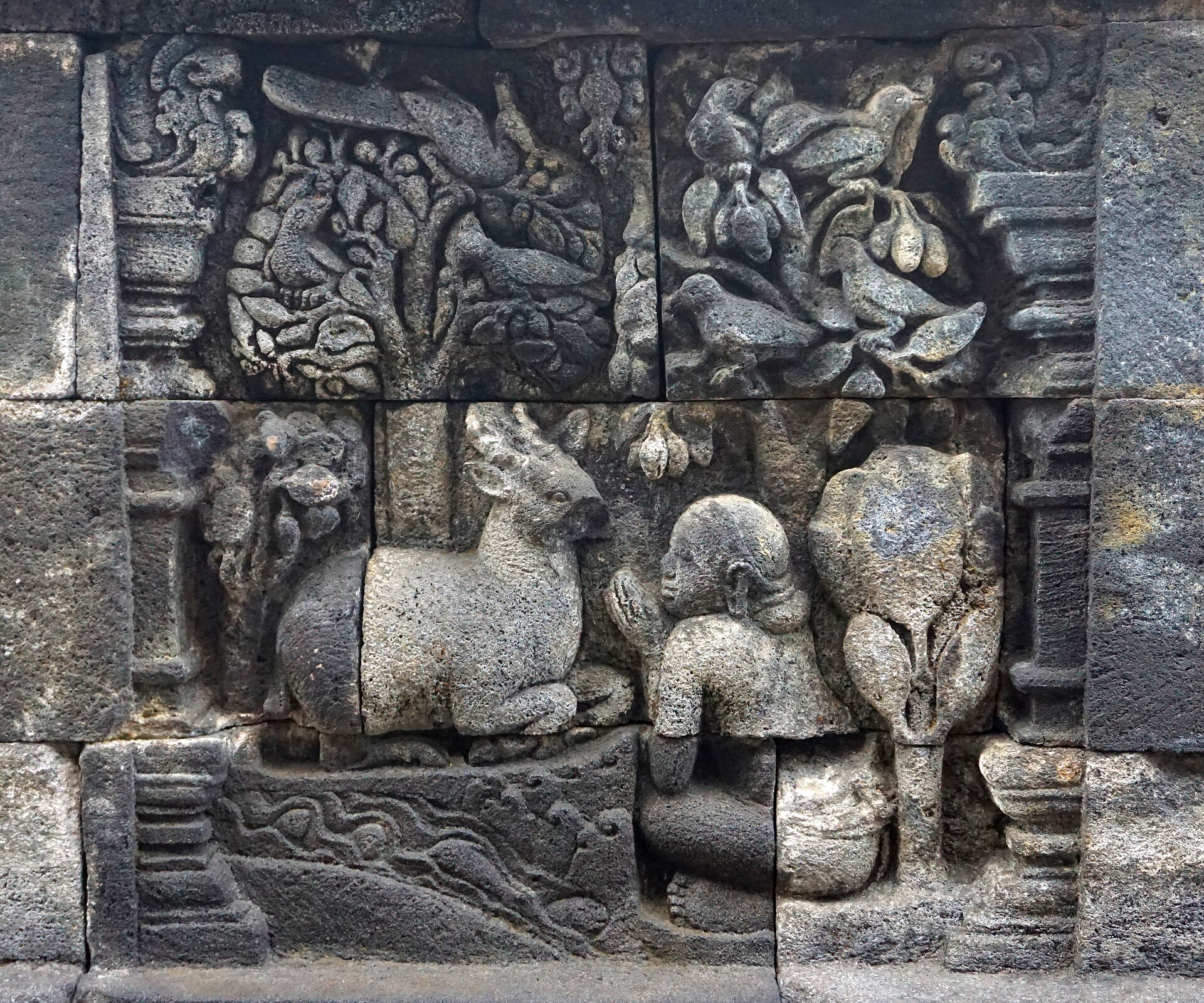 Carved Ruru deer in water beside the human it rescues, with trees and landscape details in a Borobudur Jātaka panel.