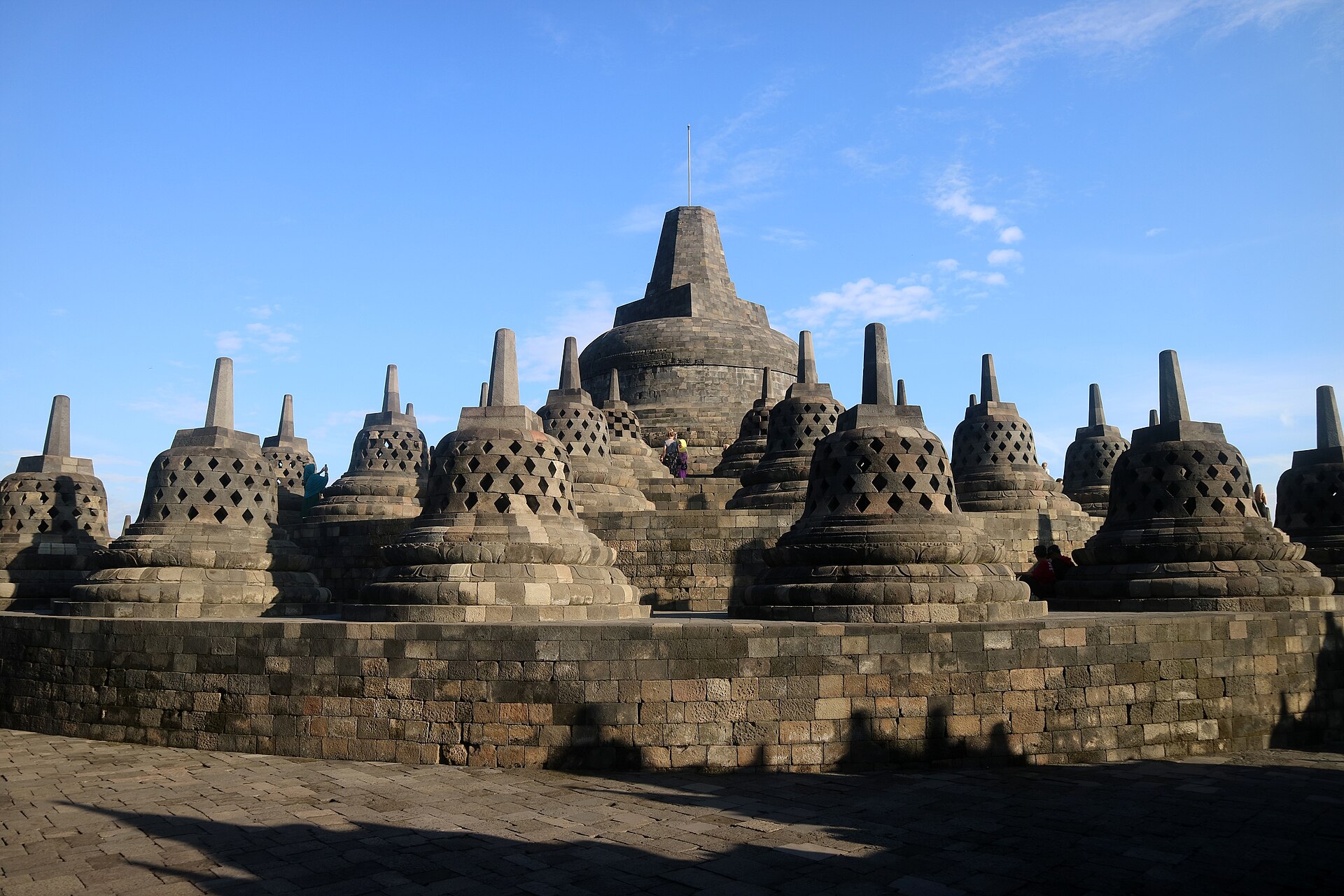Borobudur upper terrace with openwork lattice stupas flanking a large closed central stupa against sky.