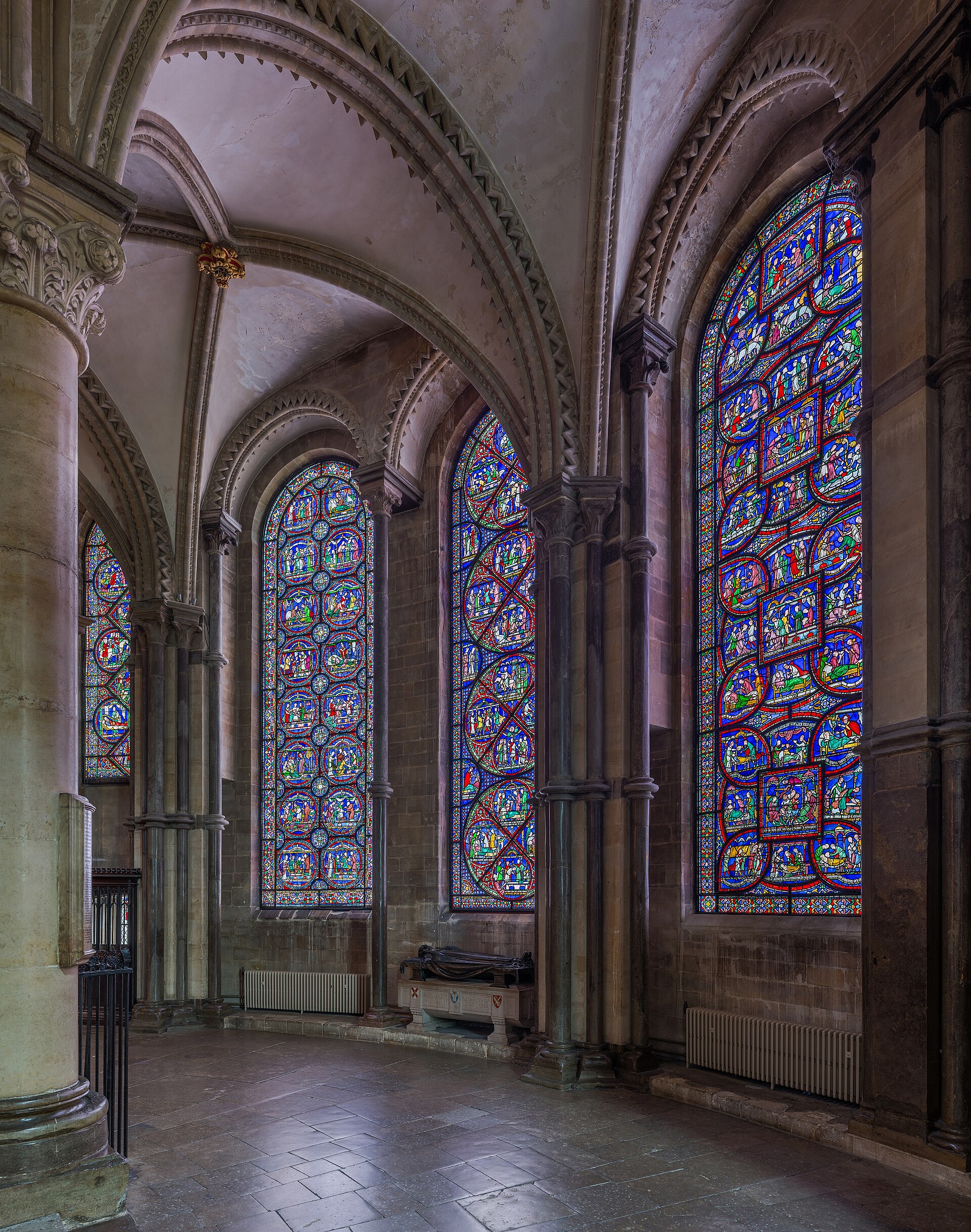 Medieval stained glass in Trinity Chapel presents small narrative scenes and standing figures within an architectural grid close to the ambulatory route.