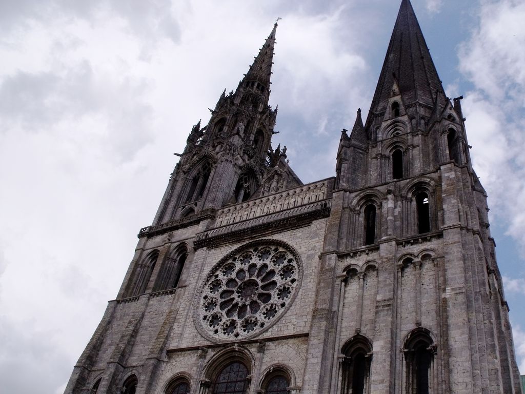 A low-angle view shows Chartres Cathedral's west front with one ornate lace-like spire, one simpler pyramidal stone spire, a rose window and dark tower openings.