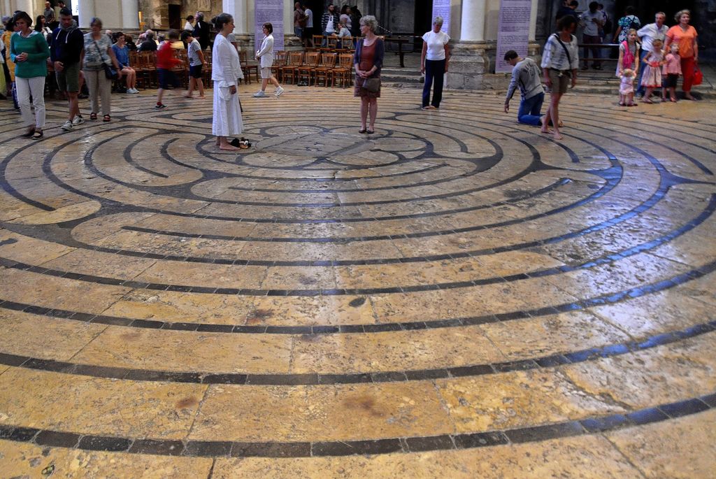 A large circular dark-stone labyrinth spreads across worn pale paving as visitors walk, stand and kneel among cathedral piers and chairs.