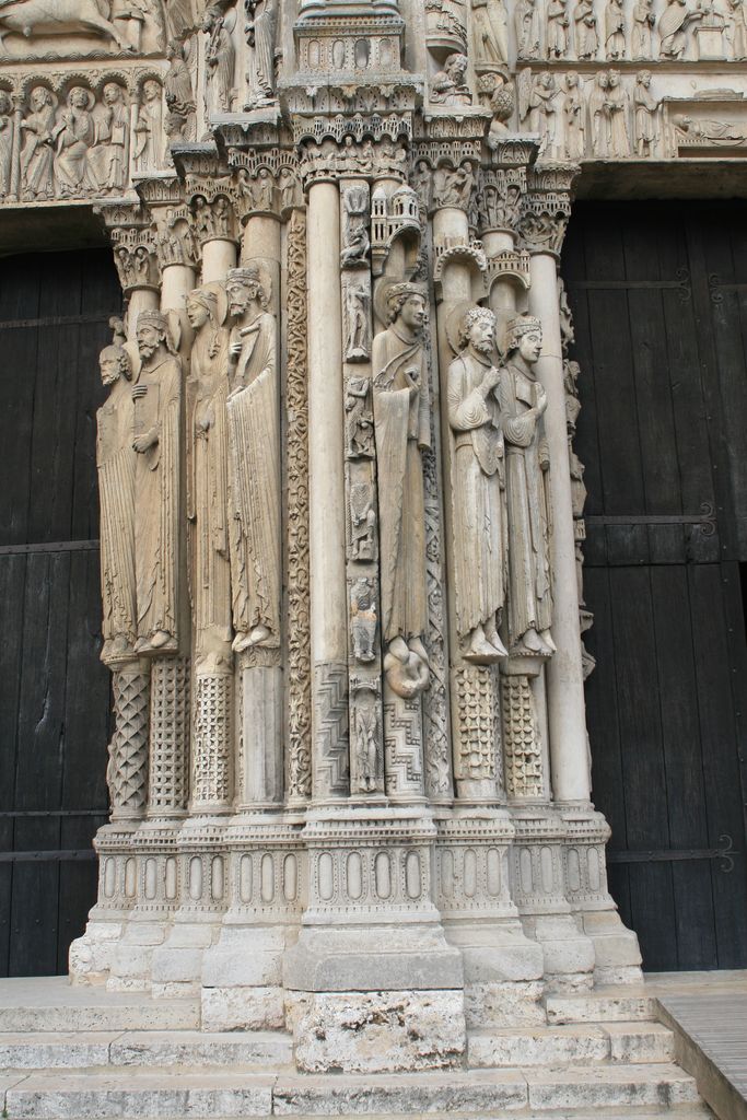Tall slender stone figures stand against carved doorway shafts beneath dense miniature canopies, between two dark wooden doors with weathered bases below.