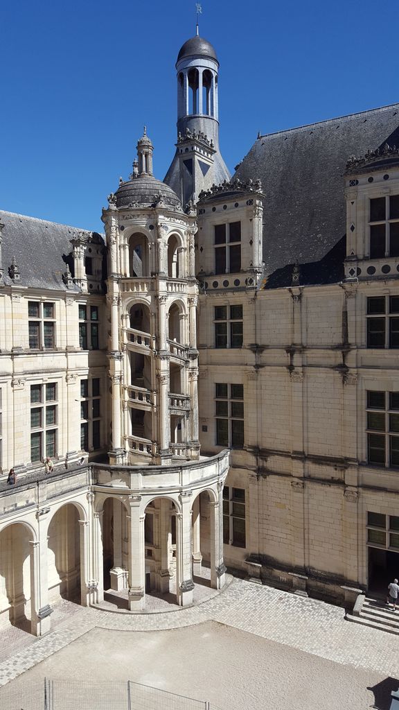 A richly carved open spiral staircase climbs an exterior courtyard turret beneath a small domed lantern beside steep roofs and regular windows.