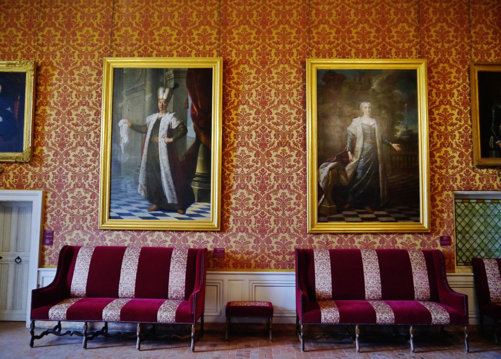 A palace room has red-and-gold patterned walls, large framed portraits, paired red settees, white panelling and a leaded window.