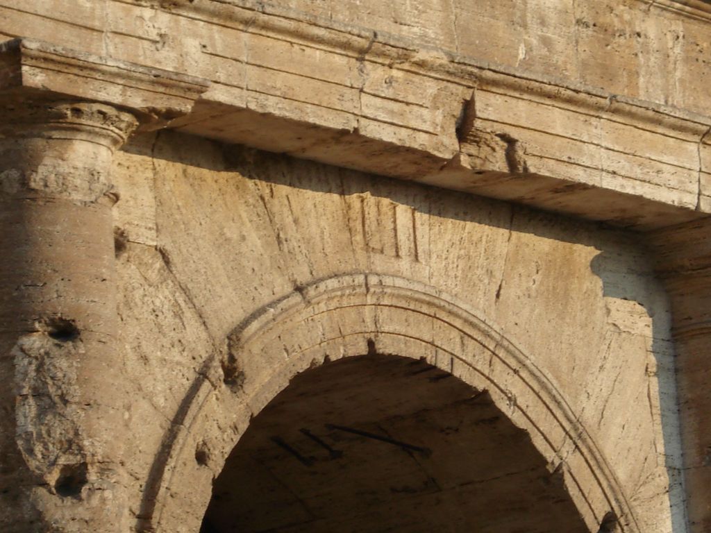 A close view of a worn travertine arch shows the Roman numeral LII carved above it, with masonry joints, damage and part of an engaged capital nearby.