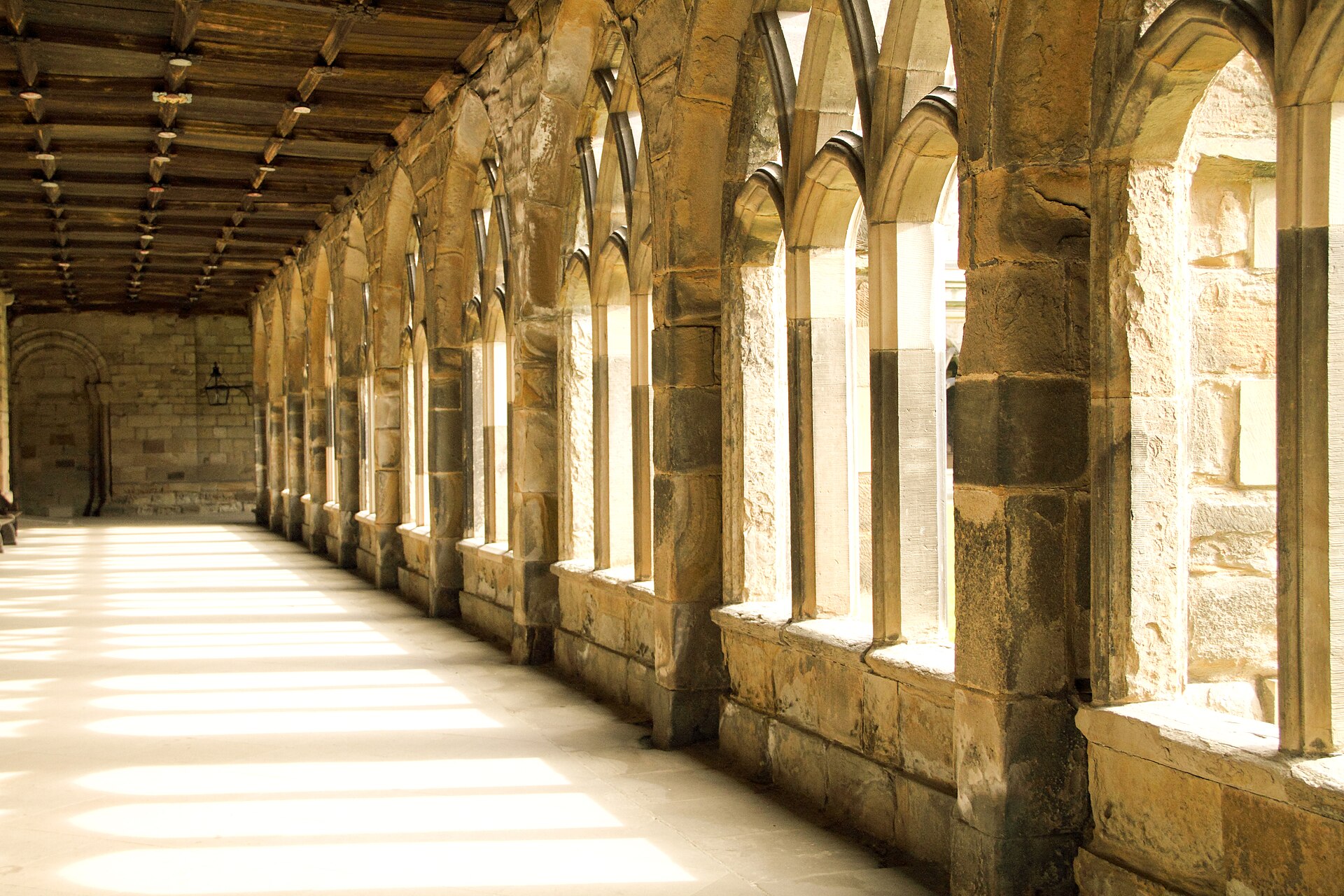 Sunlight crosses the stone paving of Durham Cathedral's cloister beside a long arcade and timber roof.