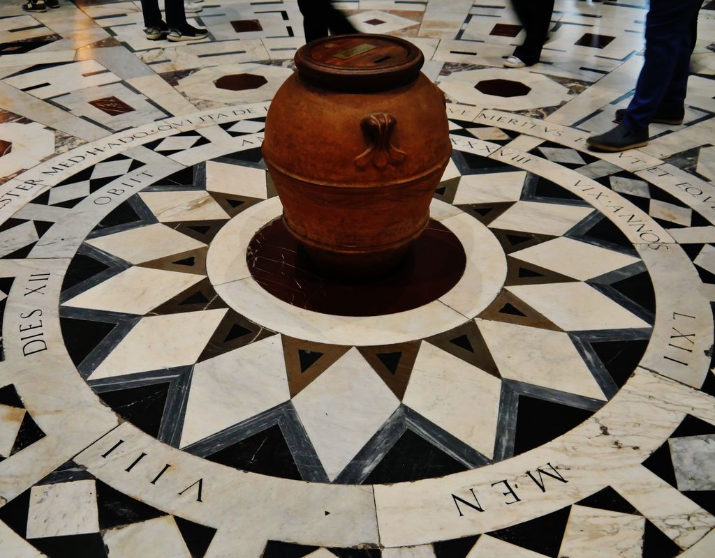 An inlaid marble floor forms concentric inscription bands, stars and polygons around a large central vessel or fixture, with visitors' feet at the edge.