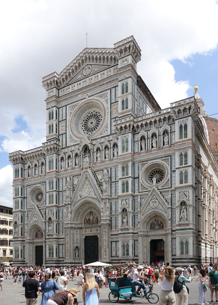 A richly patterned marble cathedral facade has three portals, many statues, three rose windows, mosaics and steep gables above a crowded square.