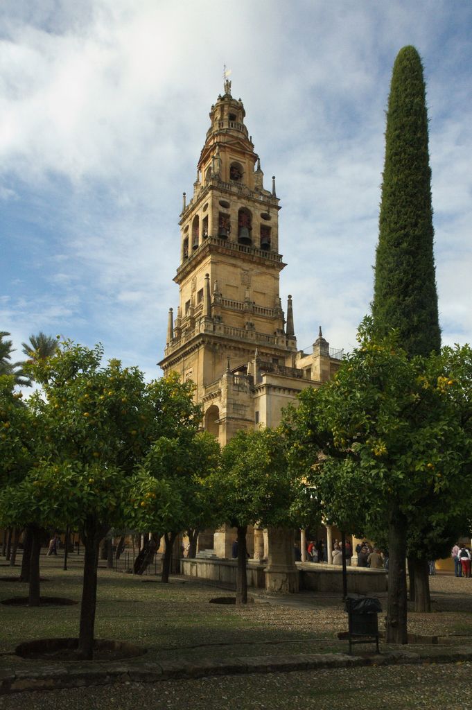 A tall tiered stone bell tower rises behind orange trees and a cypress in a sunlit courtyard with arcades and visitors.