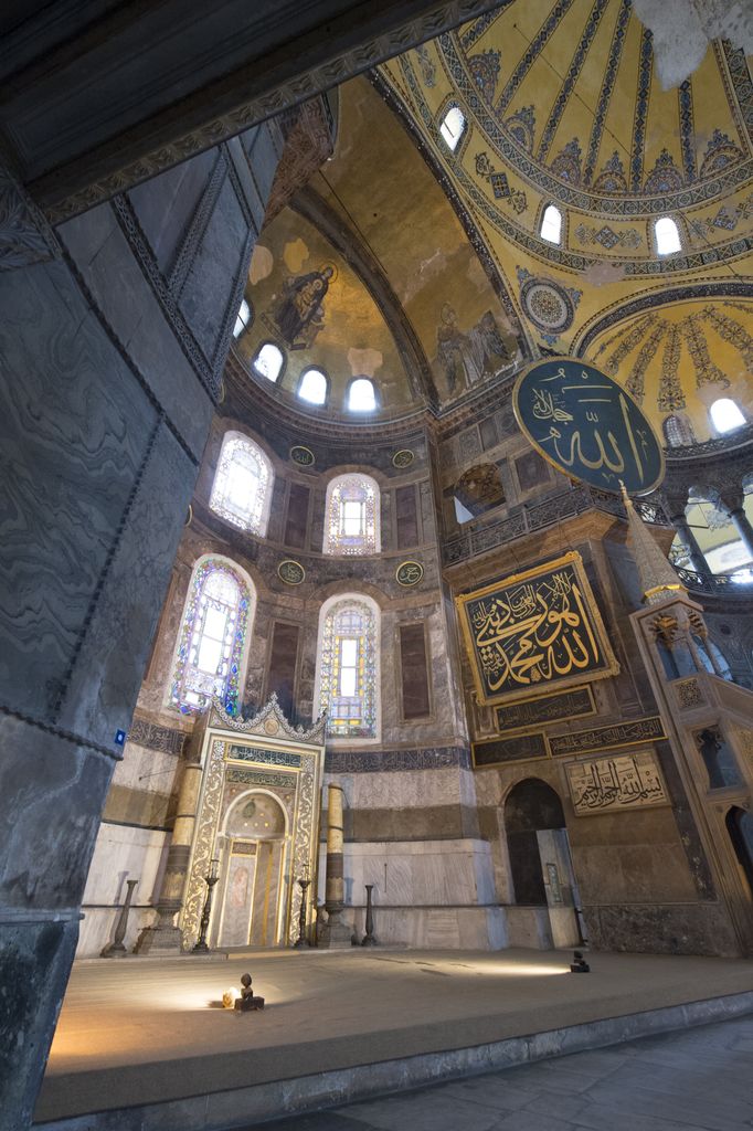 A tall decorated mihrab and large Arabic calligraphic panels occupy a marble-lined apse beneath a windowed semidome with a surviving Christian figure above.