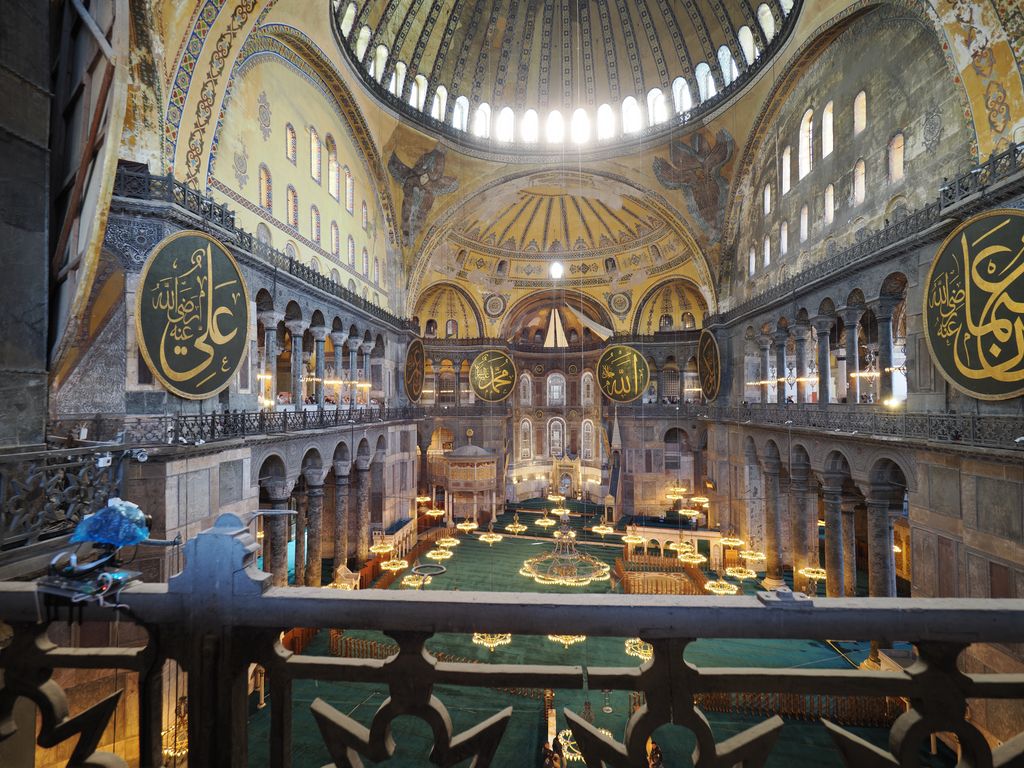 From an upper gallery, the view spans Hagia Sophia's domed nave, arcaded galleries, giant Arabic roundels, green prayer carpet, lamps, barriers and layered wall decoration.
