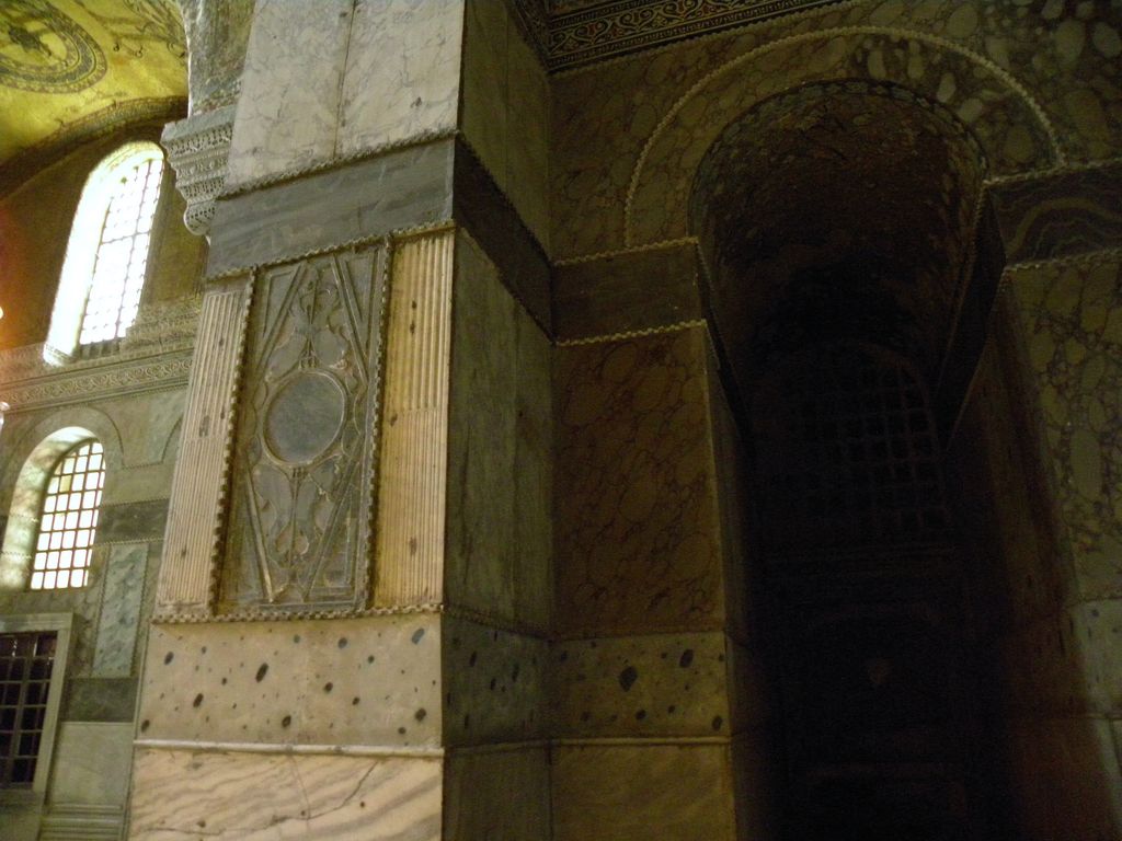 Coloured marble slabs and carved borders cover a massive interior pier and adjacent arch, with pale windows at left, surface losses and a deeply shadowed recess.