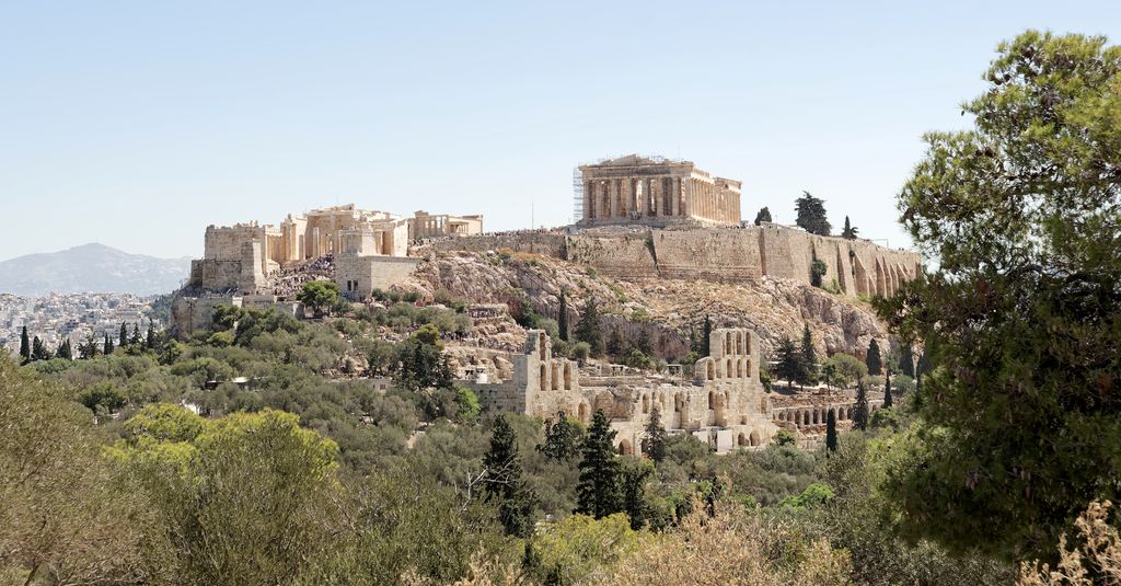 The Parthenon and other pale stone monuments stand on the rocky Acropolis above trees and Athens, with restoration scaffolding visible around the temple.