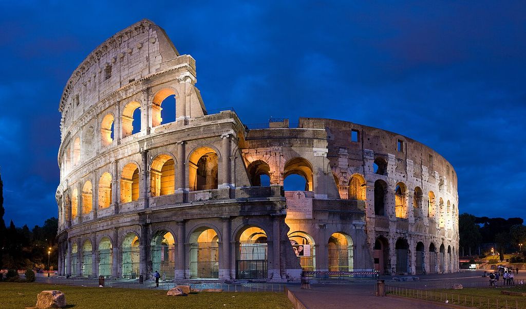 A floodlit Roman amphitheatre curves across a dusk scene, with three levels of arches, a surviving solid attic section, broken masonry and modern visitor barriers.