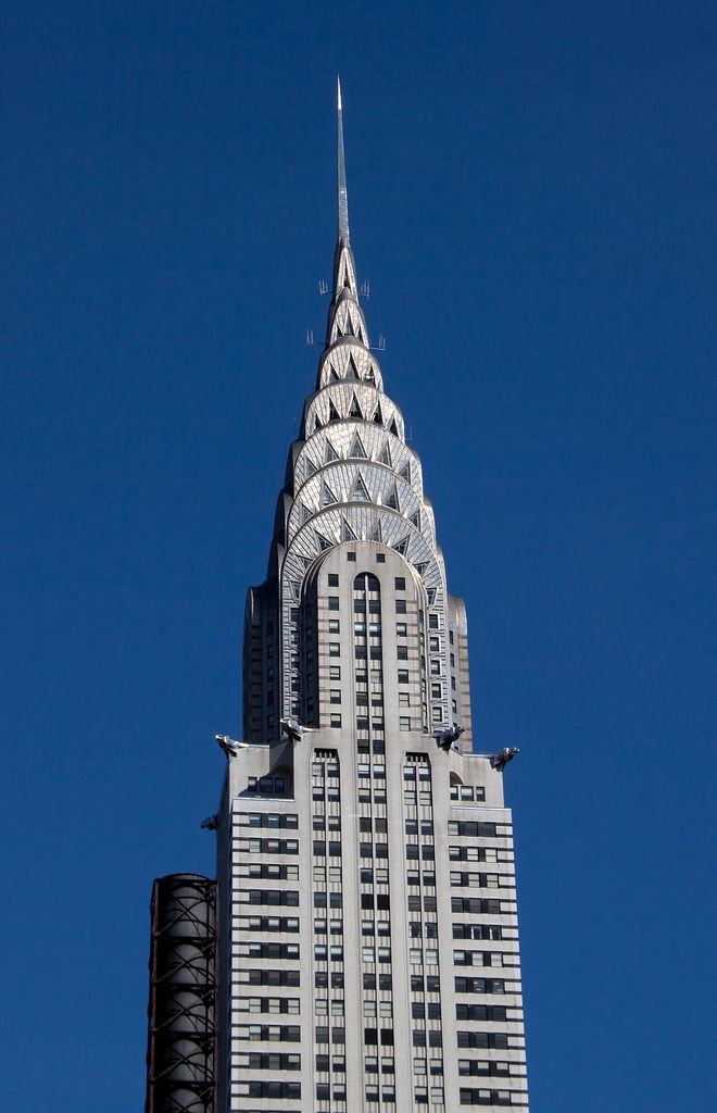 The upper Chrysler Building rises against a deep blue sky with stacked stainless-steel arches, triangular windows, eagle ornaments and a long thin spire.