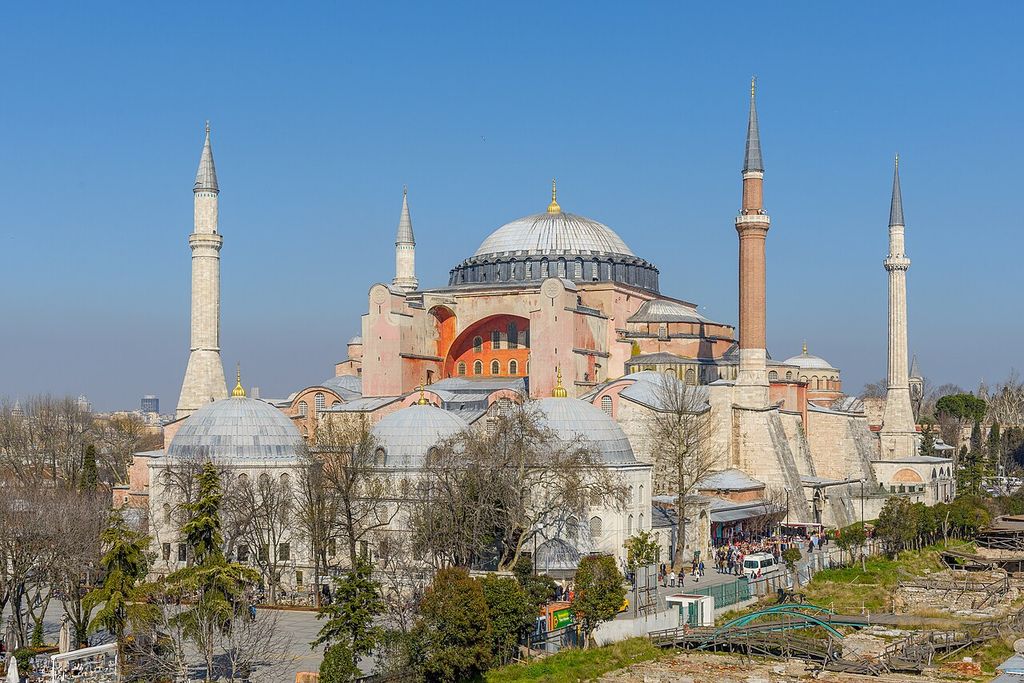 A vast pink and pale stone building rises in domes and half-domes between four slender minarets, with trees, visitors and later structures around it.
