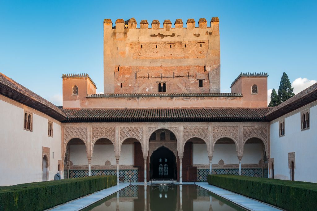 A long reflecting pool and clipped green hedges lead through a delicate white arcade to a massive reddish square tower beneath a clear blue sky.