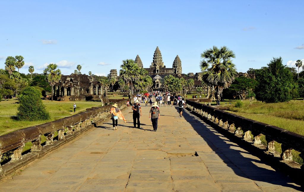 A broad stone causeway crowded with visitors runs between carved balustrades and greenery toward Angkor Wat's central towers under a bright blue sky.